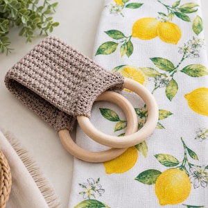 May include: A beige crocheted towel topper featuring a textured stitch pattern attached to two smooth, light-colored wooden rings, resting on a white kitchen towel decorated with a repeating pattern of yellow lemons, green leaves, and white blossoms.