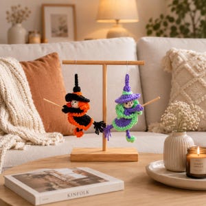 May include: Two crocheted witch ornaments hanging from a wooden T-shaped stand. One witch wears a black and orange outfit with a matching hat, while the other wears a purple and green outfit with a matching hat. Both figures hold small wooden broomsticks.