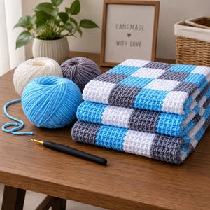 May include: A stack of three folded crochet blankets featuring a blue, white, and gray checkered pattern. Beside the blankets are three balls of yarn in matching colors and a black crochet hook with a gold tip, all resting on a wooden surface.