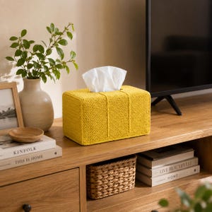 May include: A rectangular tissue box cover crafted from bright yellow yarn in a dense crochet stitch pattern. The cover features a central vertical seam detail and is designed to fit over a standard tissue box, with white tissues protruding from the top.