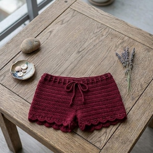 May include: Dark red crochet shorts featuring a ribbed texture, a drawstring waistband, and a scalloped hemline. The garment is displayed alongside a small ceramic dish containing several light-colored buttons and a sprig of dried lavender.