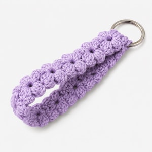 May include: A lavender crochet keychain wristlet featuring a repeating floral pattern made of yarn. The soft, textured fabric is looped into a strap and securely attached to a circular silver-toned metal split ring for holding keys.