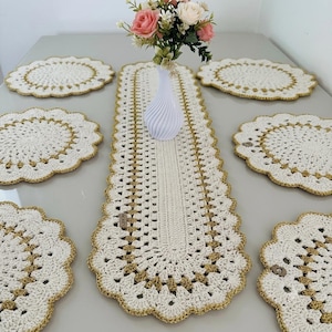 May include: A set of six round crochet placemats and one long rectangular crochet table runner, all in off-white yarn with metallic gold trim. A white vase containing pink and white artificial flowers sits on the center of the runner.