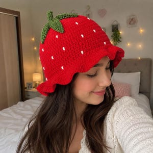 May include: A bright red crochet bucket hat shaped like a strawberry, featuring a green stem and leaves on the crown and small white embroidered dots scattered across the surface to mimic seeds, with a wavy, ruffled brim edge.