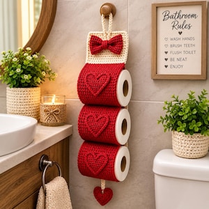 May include: A vertical crochet toilet paper holder in cream and red yarn. The holder features three red sections, each decorated with a raised red heart, and a top flap with a red bow. A small red heart hangs from the bottom of the holder.