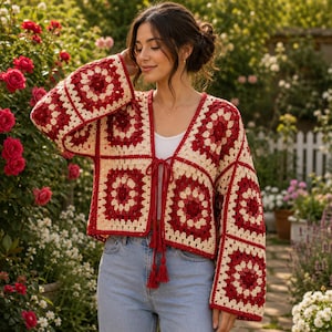 Pode incluir: Um cardigã curto feito de quadrados de crochê em fios vermelho-escuro e creme. A peça apresenta mangas longas e largas, um design frontal aberto com um fecho de laço central finalizado com duas borlas longas e um decote em V.