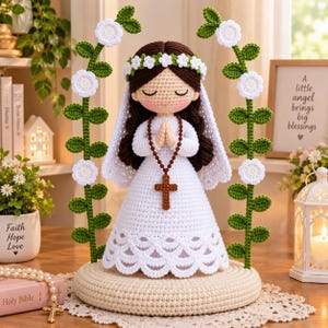 May include: A crocheted doll with brown hair, wearing a white dress and veil, adorned with a floral crown. The doll has hands clasped in prayer, holding a brown beaded rosary with a cross. It is framed by two tall, green crocheted vine structures with white flowers, all mounted on a circular base.