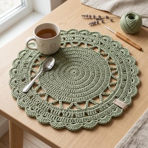 May include: A round, sage green crocheted placemat with a scalloped edge and a central circular pattern. A ceramic mug with a botanical design, a silver spoon, a ball of green yarn, and a wooden crochet hook are arranged on the placemat.
