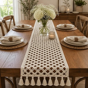 Può includere: Un runner da tavola rettangolare color crema caratterizzato da un intricato motivo a uncinetto con rombi traforati e rifinito con nappe decorative lungo i bordi. Il runner è centrato su un tavolo da pranzo in legno con coperti.