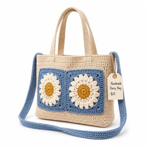 Puede incluir: Un bolso de ganchillo beige con dos parches cuadrados azules que presentan motivos de margaritas blancas y amarillas. El bolso tiene dos asas superiores y una correa larga azul. Una etiqueta de papel adjunta dice Handmade Daisy Bag.