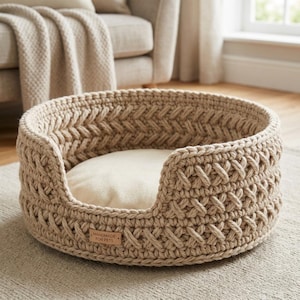 May include: A round, beige crochet pet bed with a textured, braided pattern along the exterior walls. The bed features a smooth, cream-colored interior cushion and a small, rectangular brown leather tag labeled HANDMADE FOR PETS.