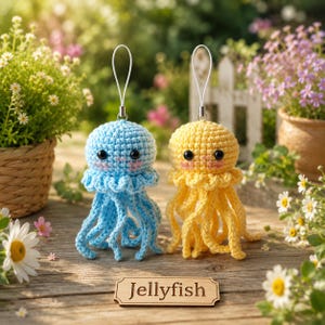 May include: Two crocheted jellyfish ornaments, one light blue and one yellow, each featuring black bead eyes, pink stitched cheeks, and a white hanging loop. A rectangular wooden sign with the word JELLYFISH is placed in front of them.