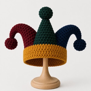 Puede incluir: Un gorro de bufón de ganchillo multicolor con una banda base de color amarillo mostaza, una punta central verde oscuro y dos puntas laterales en burdeos intenso y azul marino. Cada punta está rematada con un pompón de ganchillo a juego.