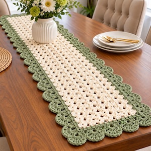 May include: A rectangular crochet table runner featuring a cream-colored center with a repeating openwork pattern, bordered by a wide, scalloped edge in sage green yarn. The runner is crafted with a textured stitch and a decorative shell border.