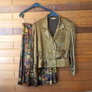 May include: A two-piece set consisting of a metallic bronze double-breasted jacket with gold-tone buttons and a matching pleated skirt featuring an abstract pattern of brown, green, and yellow blocks, both displayed on black plastic hangers.