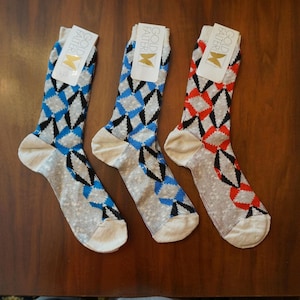 Könnte beinhalten: Drei Paar Socken mit Rautenmuster. Zwei Paare haben blaue, schwarze und graue Rauten, während ein Paar rote, schwarze und graue Rauten aufweist. Jede Socke hat ein weißes Etikett am oberen Rand mit der Aufschrift Gold Falter.