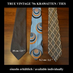 Puede incluir: Tres corbatas etiquetadas TRUE VINTAGE 70s KRAWATTEN / TIES. Una es gris oscuro liso, otra es azul claro con círculos concéntricos naranjas y negros, y otra tiene un patrón geométrico negro y crema. Anchuras de 10 cm y 8,2 cm.
