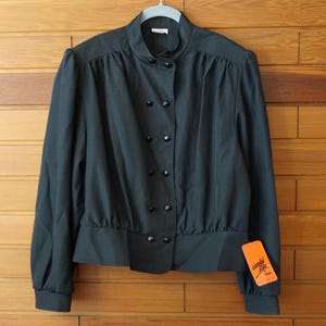 Op de afbeelding: Een donkergroene blouse met lange mouwen, een hoge kraag, een dubbele rij kleine zwarte knopen aan de voorzijde en geplooide schouders. Het kledingstuk heeft een getailleerde tailleband en een oranje label met de tekst Combi Life.