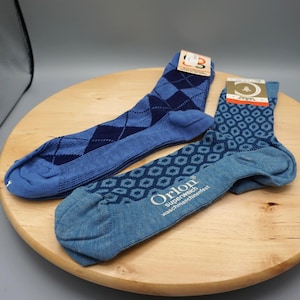 Op de afbeelding: Twee paar blauwe sokken met labels. Eén paar heeft een donkerblauw ruitpatroon op een lichtblauwe achtergrond. Het andere paar heeft een geometrisch patroon en bevat de tekst Orlon superweich waschmaschinenfest aan de zijkant.