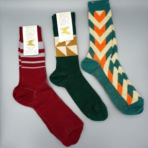 Könnte beinhalten: Drei Paar Socken mit Etiketten der Marke Gold Falter. Das erste Paar ist kastanienbraun mit grauen Streifen. Das zweite ist dunkelgrün mit einem goldenen und weißen geometrischen Muster am Schaft. Das dritte zeigt ein Zickzackmuster in Petrol, Orange und Creme.