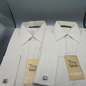 May include: Two long-sleeved button-down shirts in a light beige fabric with thin vertical stripes. Each shirt features a pointed collar, a gold-colored tag labeled Frisa Wäsche Baumwolle, and a decorative cufflink attached to the sleeve cuff.
