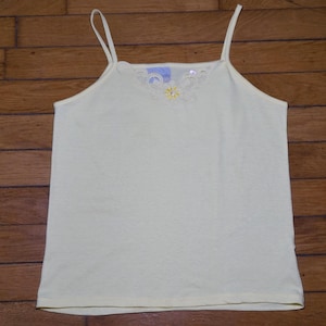 May include: A pale yellow camisole featuring thin spaghetti straps and a square neckline. The upper chest area is decorated with a delicate, tonal embroidered floral pattern that includes a small yellow flower center with a subtle rhinestone accent.