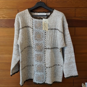 Könnte beinhalten: Ein langärmeliger Strickpullover in Beige und Hellgrau mit einem zentralen vertikalen Einsatz aus fünf quadratischen Häkelmotiven. Das Kleidungsstück hat einen dunkelbraunen, gewellten Saum am Ausschnitt, an den Ärmelbündchen und am Saum.
