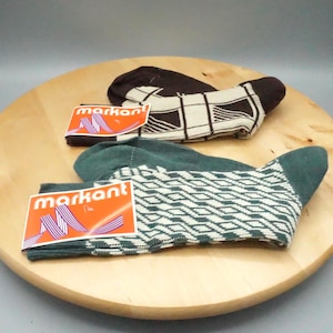 Op de afbeelding: Twee paar sokken met aangehechte labels met de tekst markant. Eén paar is donkerbruin met een crèmekleurig ruitpatroon. Het andere paar is donkergroen met een crèmekleurig diamantpatroon. De labels zijn oranje met witte tekst en een paars lint.