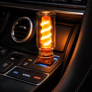May include: A cylindrical car accessory with a clear glass outer shell and an internal orange core featuring a glowing white spiral filament. The device is plugged into a vehicle console near buttons labeled ECO, NORMAL, and SPORT.
