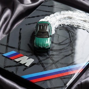 Pode incluir: Um carro de brinquedo verde montado numa placa retangular preta brilhante. A placa apresenta o logótipo M, uma faixa tricolor em azul, roxo e vermelho, e marcas de pneus pretas. Um material branco texturado simula um rasto de fumo.
