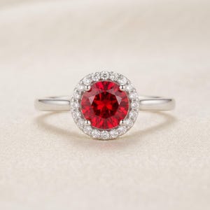 May include: A silver-toned ring featuring a round, vibrant red centre stone surrounded by a halo of small, clear, sparkling stones. The band is smooth and polished, leading up to a four-prong setting that secures the central red gemstone.