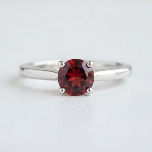 May include: A silver ring featuring a round, faceted deep red garnet gemstone set in a four-prong mounting. The band is smooth, polished, and metallic, showcasing a classic design with a central stone that displays a vibrant, dark crimson hue.