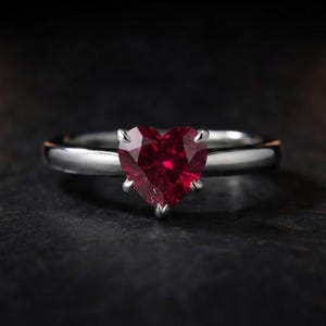 Op de afbeelding: Een zilverkleurige ring met een gefacetteerde, hartvormige rode edelsteen in een zetting met vier pootjes. De ringband is glad en gepolijst en heeft een strakke metalen afwerking die contrasteert met de dieprode kleur van de hartvormige steen.