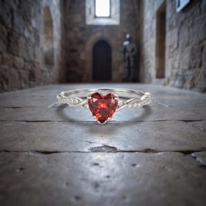 May include: A silver-toned ring featuring a heart-shaped red gemstone at the center, flanked by small clear stones set along a twisted band. The metal band has a polished finish and the design incorporates a delicate, interwoven structure.