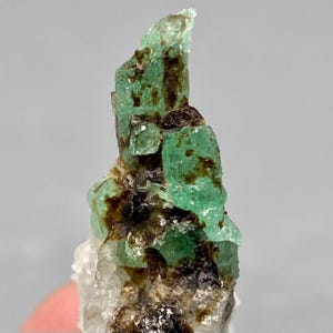 以下が含まれることがあります： 半透明で鮮やかな緑色の結晶が、暗褐色の土のような内包物と共に集まった原石の標本。構造はギザギザとして不規則であり、結晶の表面全体にエメラルドグリーンと深い茶色の様々な色合いが見られる、自然で磨かれていない質感が特徴です。