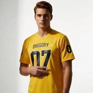 Op de afbeelding: Een geel T-shirt met V-hals voorzien van de tekst DIGGORY in het zwart, het getal 07 in grote zwarte cijfers en het woord HUFFLEPUFF in kleinere zwarte letters. De mouwen zijn voorzien van het cijfer 0 in het zwart en een wit dassymbool.