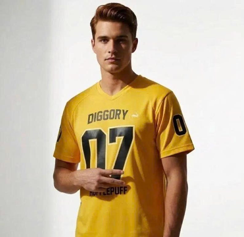 Op de afbeelding: Een geel T-shirt met V-hals voorzien van de tekst DIGGORY in het zwart, het getal 07 in grote zwarte cijfers en het woord HUFFLEPUFF in kleinere zwarte letters. De mouwen zijn voorzien van het cijfer 0 in het zwart en een wit dassymbool.