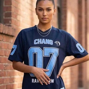 Op de afbeelding: Een donkerblauw shirt met korte mouwen, voorzien van de tekst CHANG en het nummer 07 in lichtblauw met witte omlijning. Het shirt bevat een kleine vogelafbeelding, de tekst RAVENCLAW, het nummer 07 op de mouwen en een wit label aan de zoom.