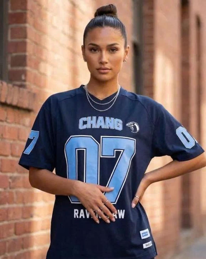 Op de afbeelding: Een donkerblauw shirt met korte mouwen, voorzien van de tekst CHANG en het nummer 07 in lichtblauw met witte omlijning. Het shirt bevat een kleine vogelafbeelding, de tekst RAVENCLAW, het nummer 07 op de mouwen en een wit label aan de zoom.