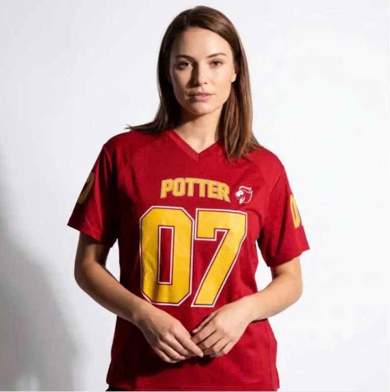 Op de afbeelding: Een rood shirt met V-hals en korte mouwen met het woord POTTER in gele letters boven het grote gele nummer 07. Een klein geel leeuwembleem bevindt zich rechts van de tekst. Het nummer 07 staat ook op elke mouw gedrukt.
