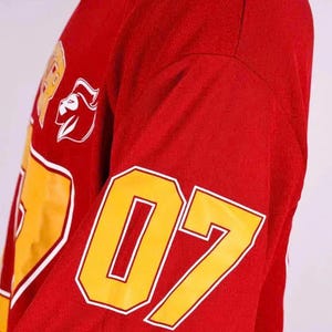 Op de afbeelding: Een rood shirt met lange mouwen en een groot geel nummer 07 op de mouw. De voorkant van het shirt toont een gele afbeelding met een witte omlijning en een wit logo van een gestileerde leeuwenkop, samen met gedeeltelijke gele tekst.