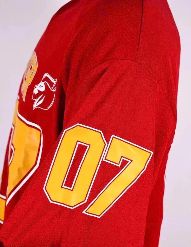Op de afbeelding: Een rood shirt met lange mouwen en een groot geel nummer 07 op de mouw. De voorkant van het shirt toont een gele afbeelding met een witte omlijning en een wit logo van een gestileerde leeuwenkop, samen met gedeeltelijke gele tekst.