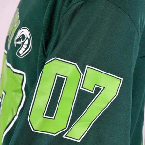 Op de afbeelding: Een donkergroen shirt met lange mouwen voorzien van een groot limoengroen nummer 07 met een witte omlijning op de mouw. Een witte afbeelding van een slangenkop is zichtbaar op het bovenste borstgedeelte, tegen de donkergroene stof.