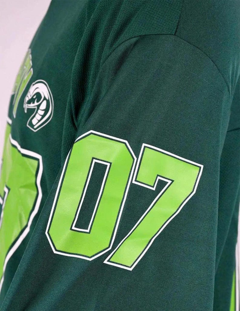 Op de afbeelding: Een donkergroen shirt met lange mouwen voorzien van een groot limoengroen nummer 07 met een witte omlijning op de mouw. Een witte afbeelding van een slangenkop is zichtbaar op het bovenste borstgedeelte, tegen de donkergroene stof.