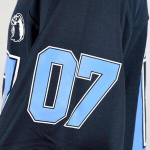 Op de afbeelding: Een donker marineblauw shirt met korte mouwen voorzien van het nummer 07 in lichtblauw met een witte omlijning op de mouw. Een wit logo van een vogelkop is geplaatst boven het nummer op het schoudergedeelte van het sportkledingstuk.