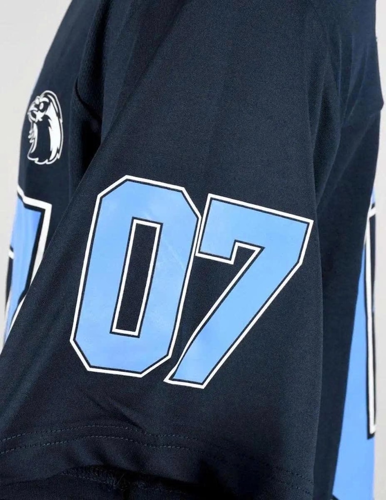 Op de afbeelding: Een donker marineblauw shirt met korte mouwen voorzien van het nummer 07 in lichtblauw met een witte omlijning op de mouw. Een wit logo van een vogelkop is geplaatst boven het nummer op het schoudergedeelte van het sportkledingstuk.