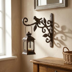 May include: A dark brown metal wall-mounted bracket featuring a decorative scrollwork design with leaf accents and a small bird figure. A matching dark brown metal lantern with glass panels and a small candle inside hangs from the bracket hook.