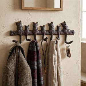May include: A dark brown cast iron wall-mounted hook rack featuring seven cat-shaped silhouettes. The tails of the cats form the hooks, which hold a tweed jacket, a plaid scarf, a braided leash, a beige scarf, and a set of metal keys on a ring.