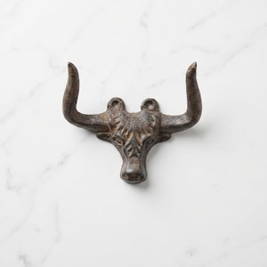 May include: A dark brown, cast iron wall hook shaped like a bull head with prominent horns. The metal surface features a textured, weathered finish with visible detailing on the forehead and snout. Two circular mounting holes are located at the top.
