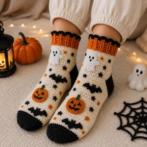 Puede incluir: Un par de calcetines de ganchillo color crema con temática de Halloween, con motivos de calabazas naranjas, fantasmas blancos y murciélagos negros. Tienen un puño naranja acanalado con borde festoneado negro y puntera y talón negros.
